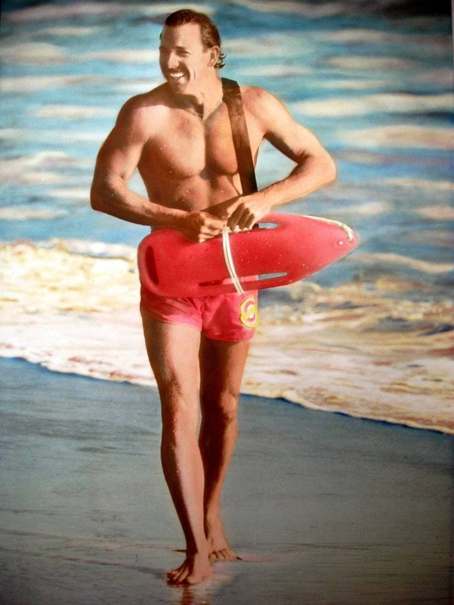 A smiling lifeguard in red shorts walks out of the surf carrying a red rescue can