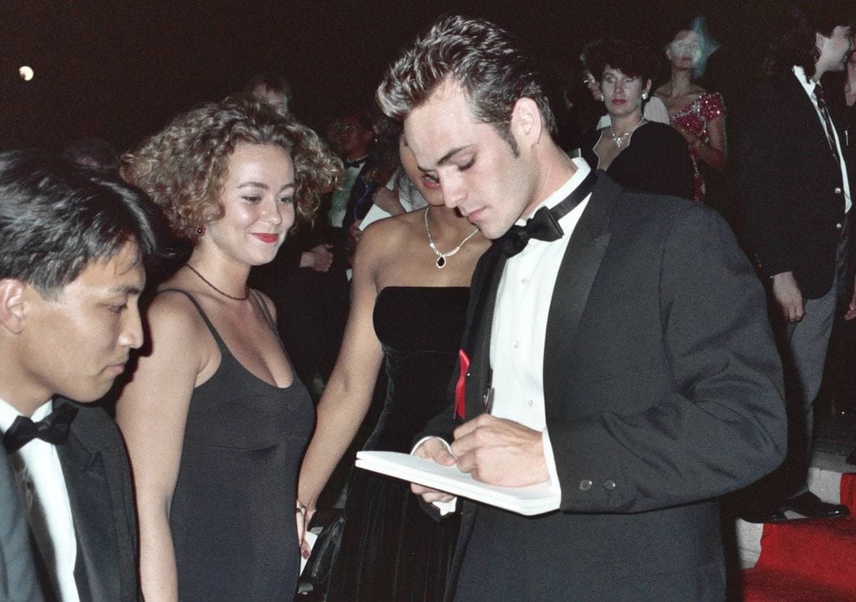 Luke Perry in a dark jacket leaning in to sign an autograph in a crowd