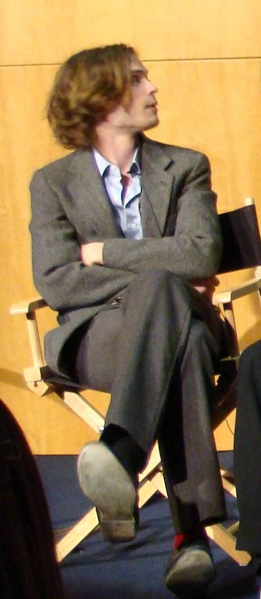 A slim young man with tousled hair in a dark jacket