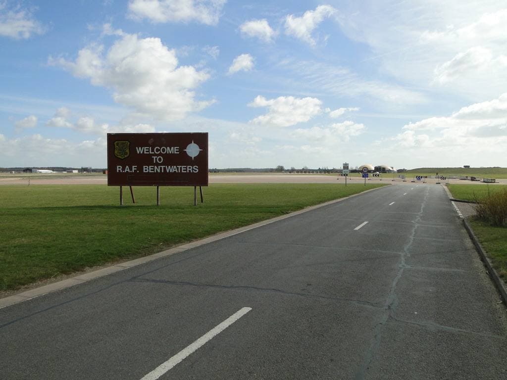 A straight empty road across grass towards a sign welcoming visitors to RAF Bentwaters