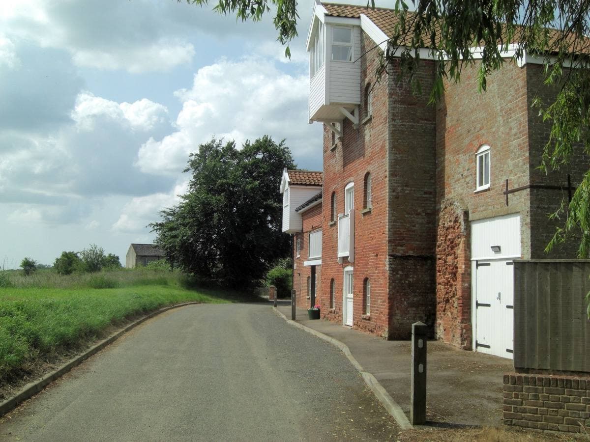 A narrow lane curving past a tall brick mill building with a white weatherboarded hoist