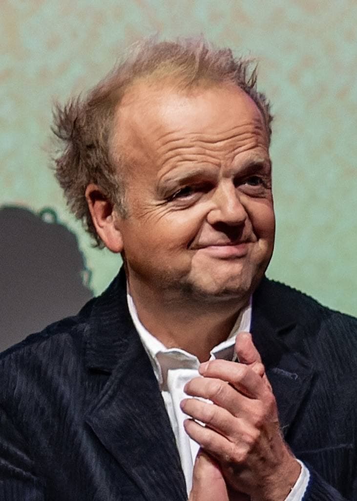 A balding man in a dark velvet jacket, hands clasped, smiling at an evening event