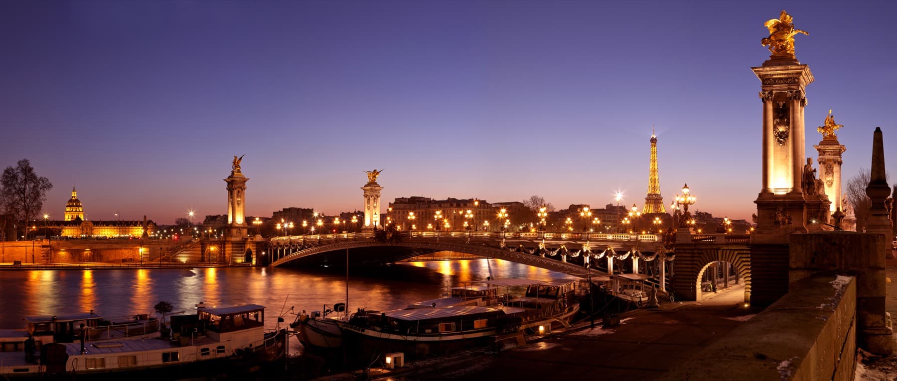 The Seine at dusk with a lit stone bridge, gilded statues on columns and the Eiffel Tower in the distance