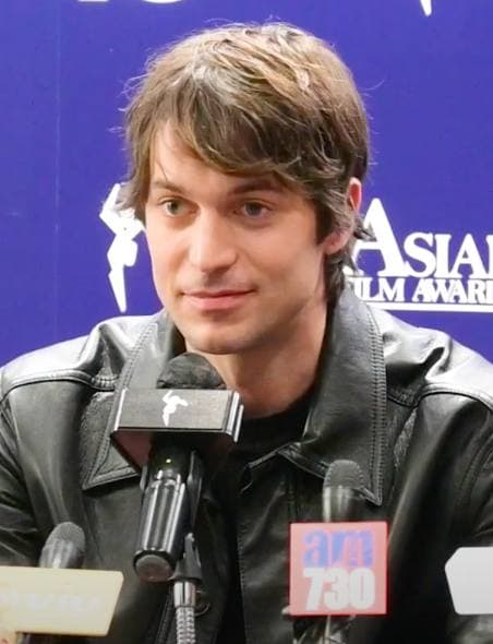 Lucas Bravo with tousled brown hair in a black leather jacket, at a microphone