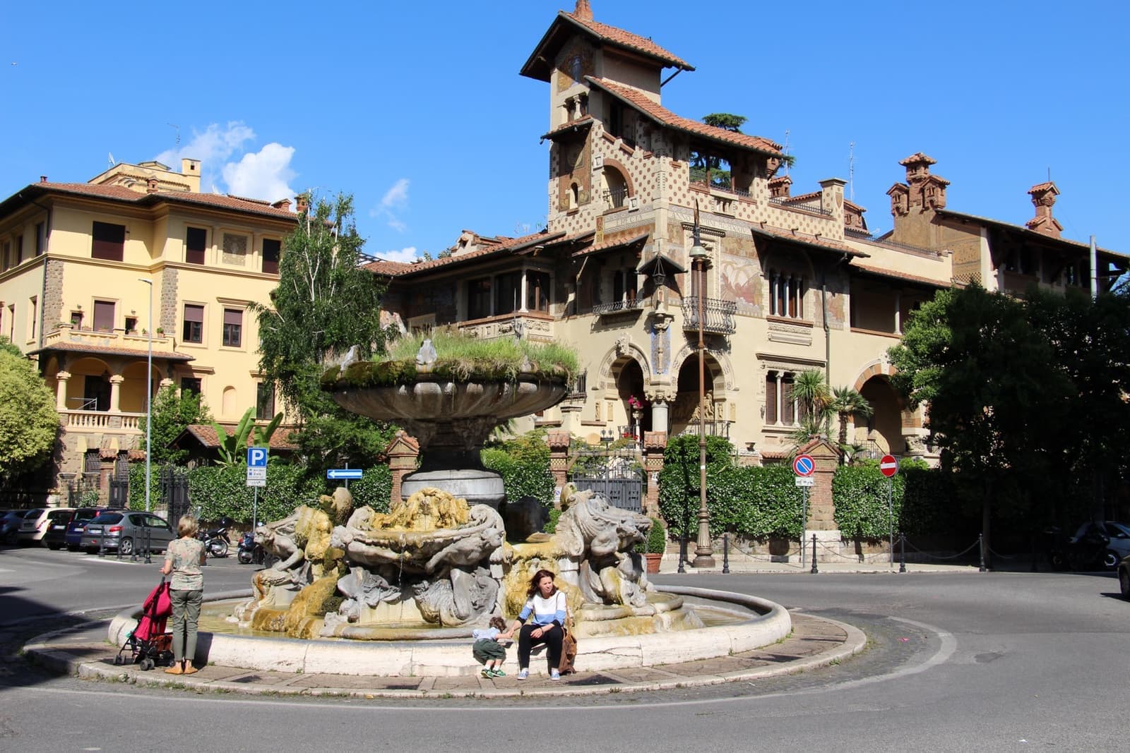 A stone fountain with frogs in a Roman piazza, ornate turreted villas behind it