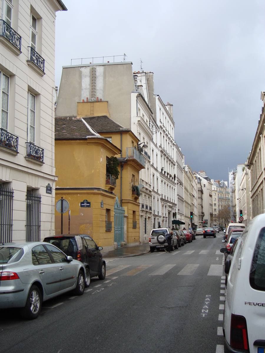 A narrow Paris street of pale apartment buildings and one ochre house, cars parked along one side