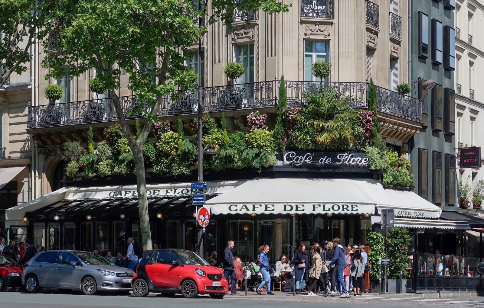 A Paris corner café with a white awning, green plants on the balconies and a crowded terrace