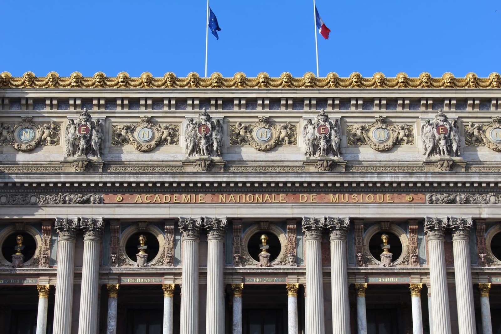 The gilded front of the Paris opera house under a blue sky, columns and statues along its facade