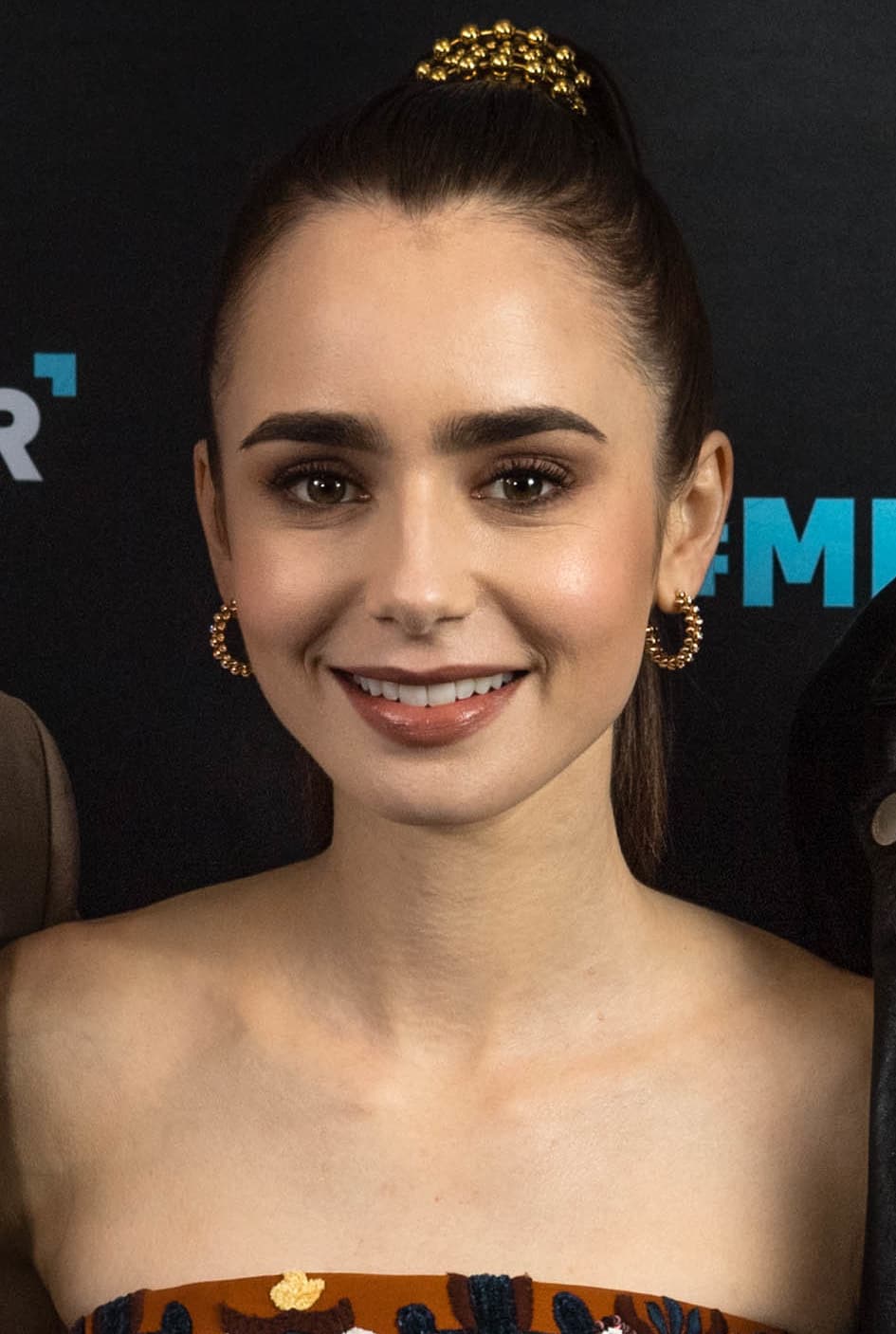 Lily Collins smiling, dark hair swept up under a gold hair comb, gold earrings