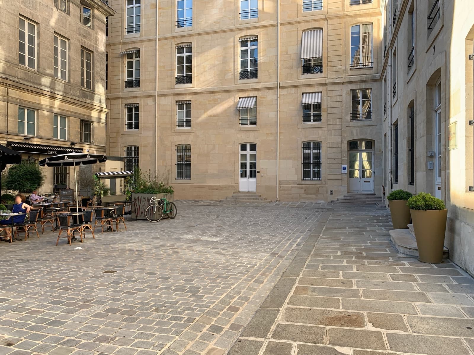 A quiet paved square of tall stone buildings with café tables and potted shrubs