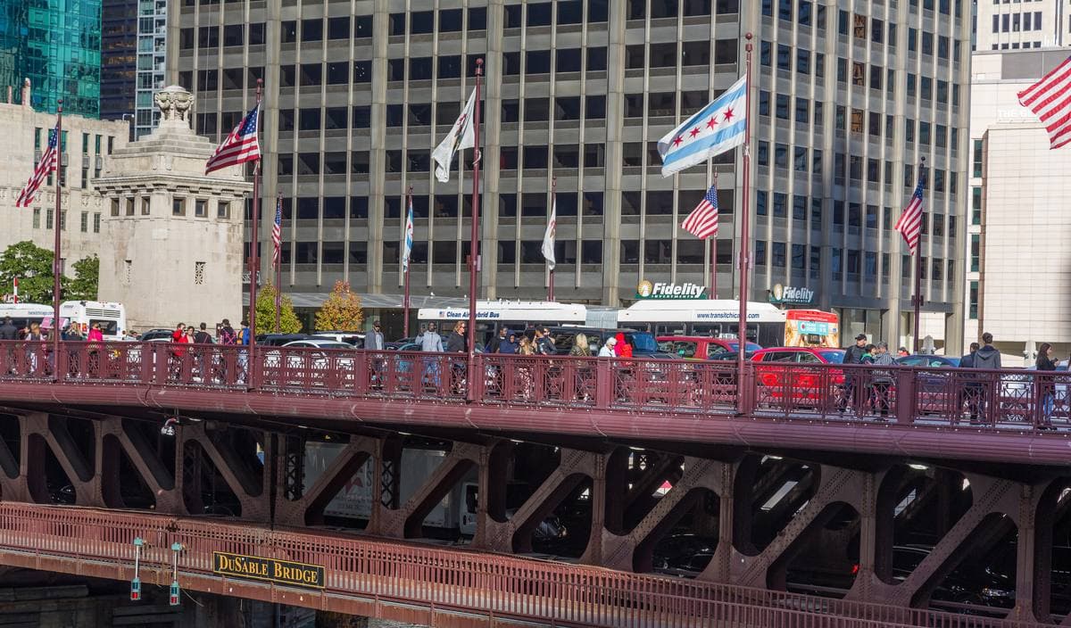 A red-painted steel bascule bridge lined with flags and people, office towers behind