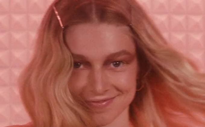Hunter Schafer smiling, long fair hair, against a pink wall