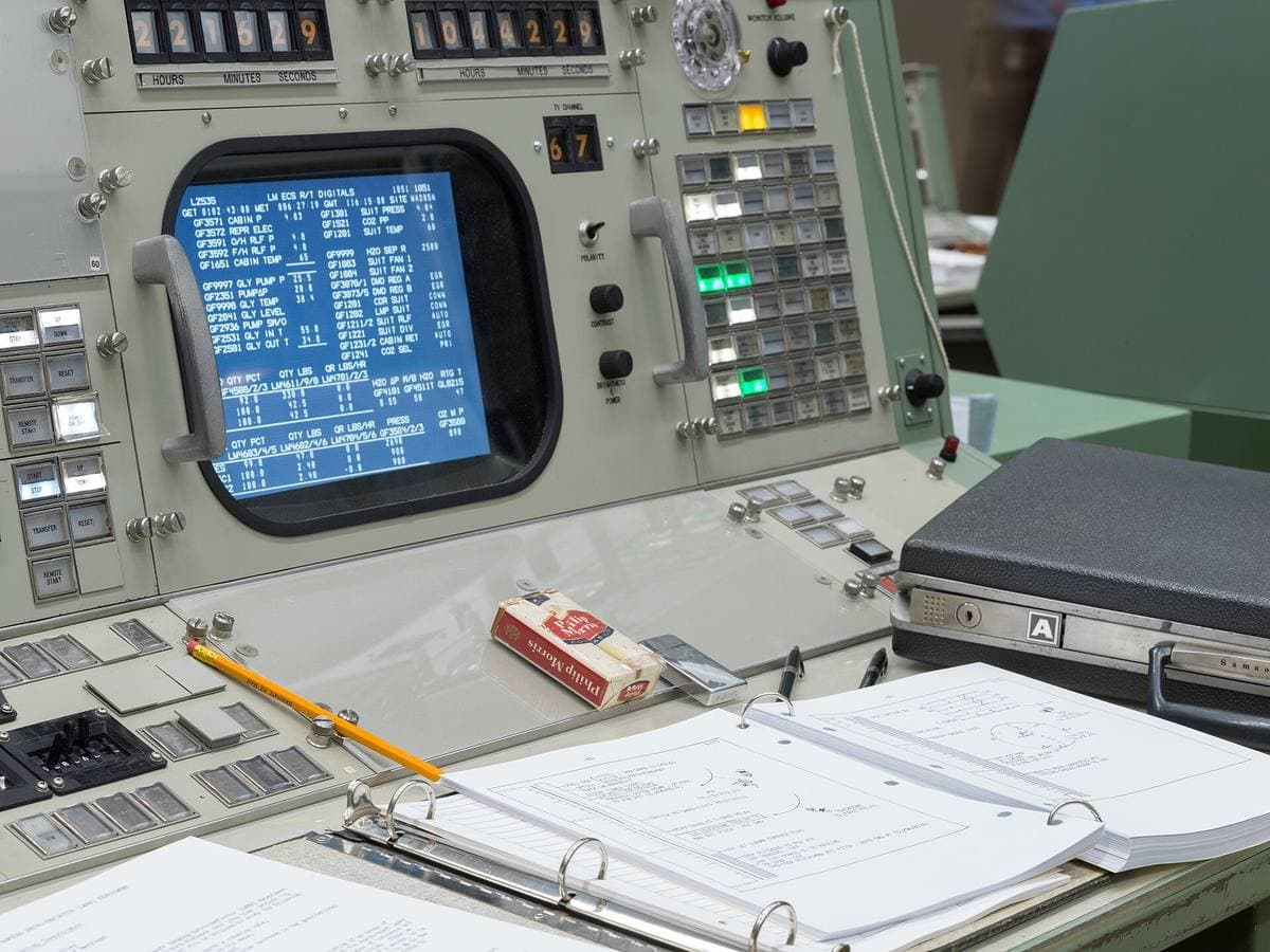 Rows of two-tone green Mission Control consoles with papers, headsets and ashtrays on the desks
