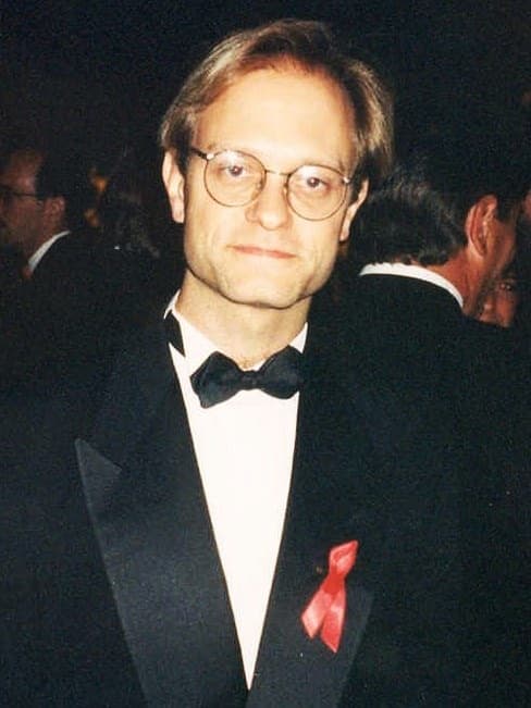 David Hyde Pierce in round glasses and black tie with a red ribbon