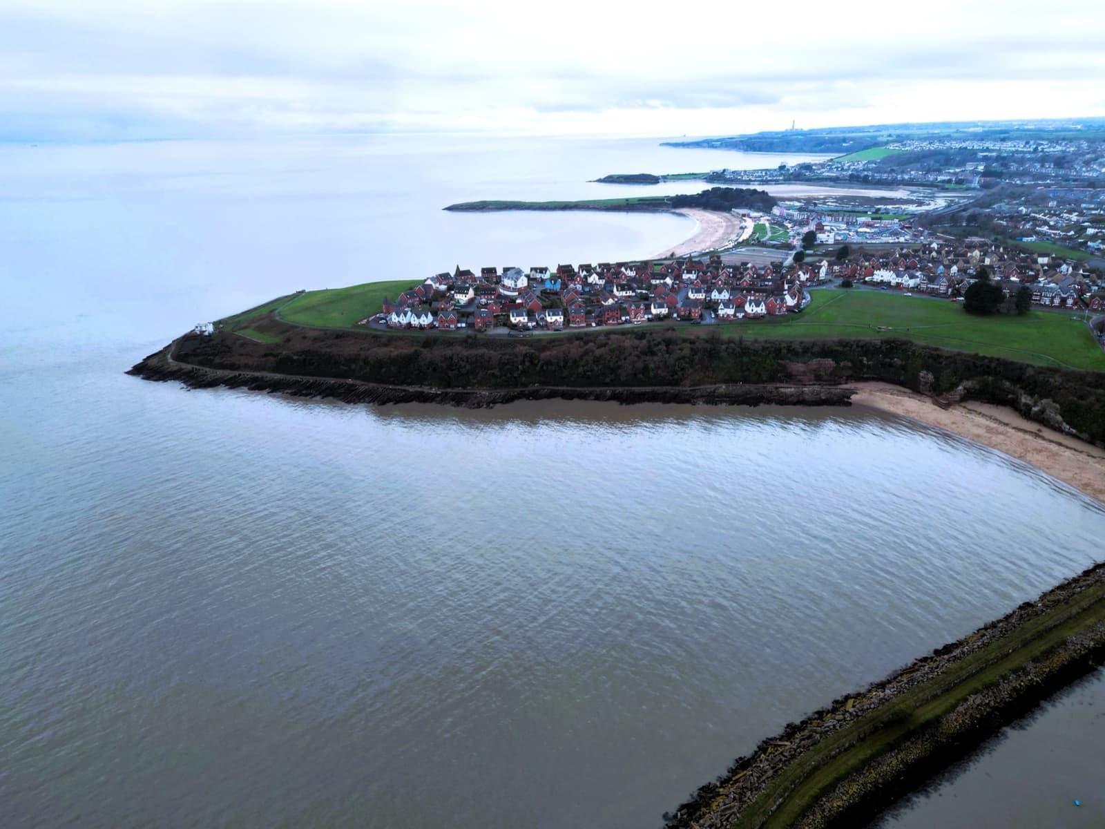 A green headland of houses ringed by grey sea, with the curve of a beach beyond