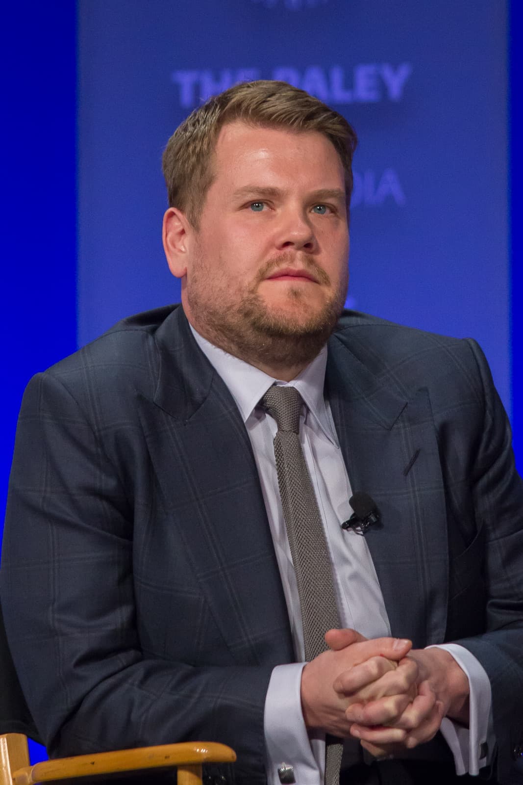 James Corden in a grey suit and tie, seated, hands clasped