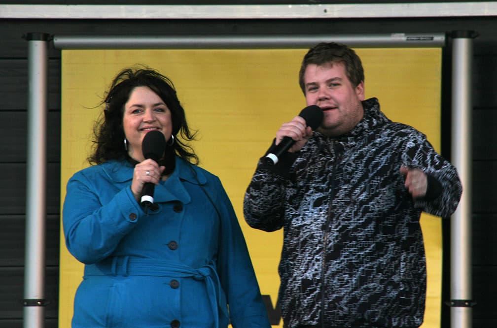 Ruth Jones in a blue coat and James Corden in a patterned jacket, both holding microphones, laughing on an outdoor stage