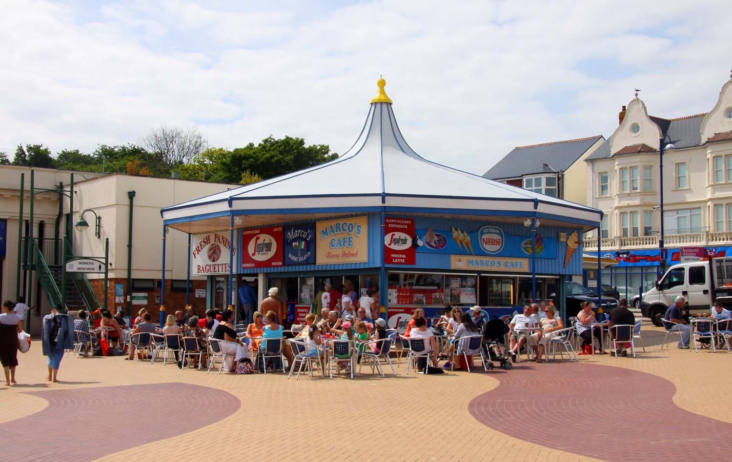 A round blue café kiosk with a peaked white roof, its terrace full of people