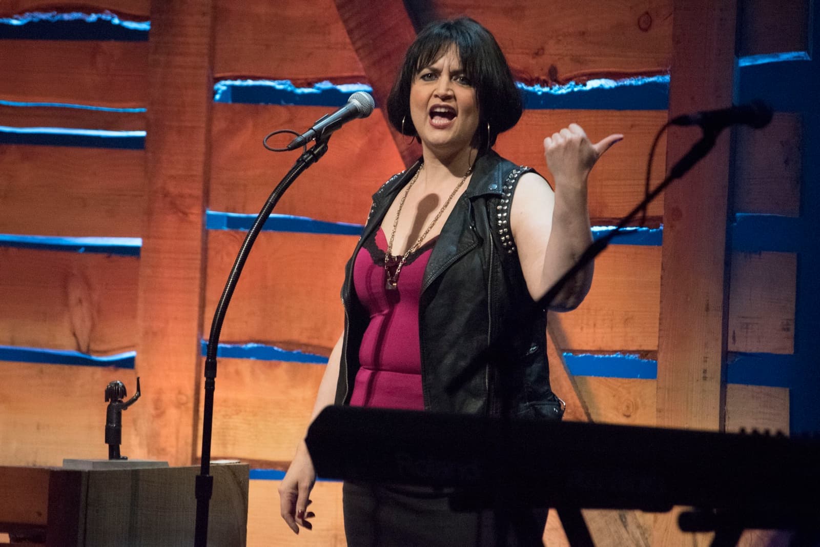 Ruth Jones in a black studded waistcoat at a microphone, thumb jerked over her shoulder