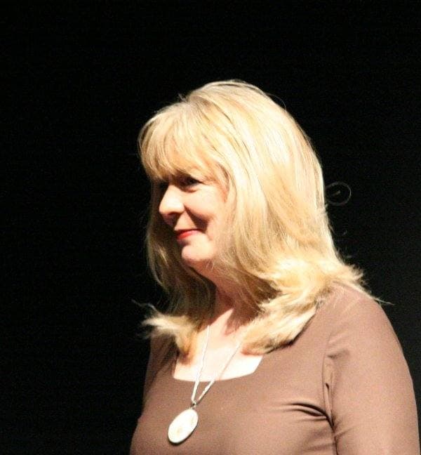 Alison Steadman with blonde hair and a brown top, holding a script on a dark stage