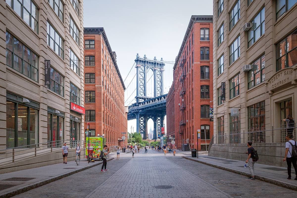 A cobbled Dumbo street between brick warehouses, the Manhattan Bridge tower framed at its end