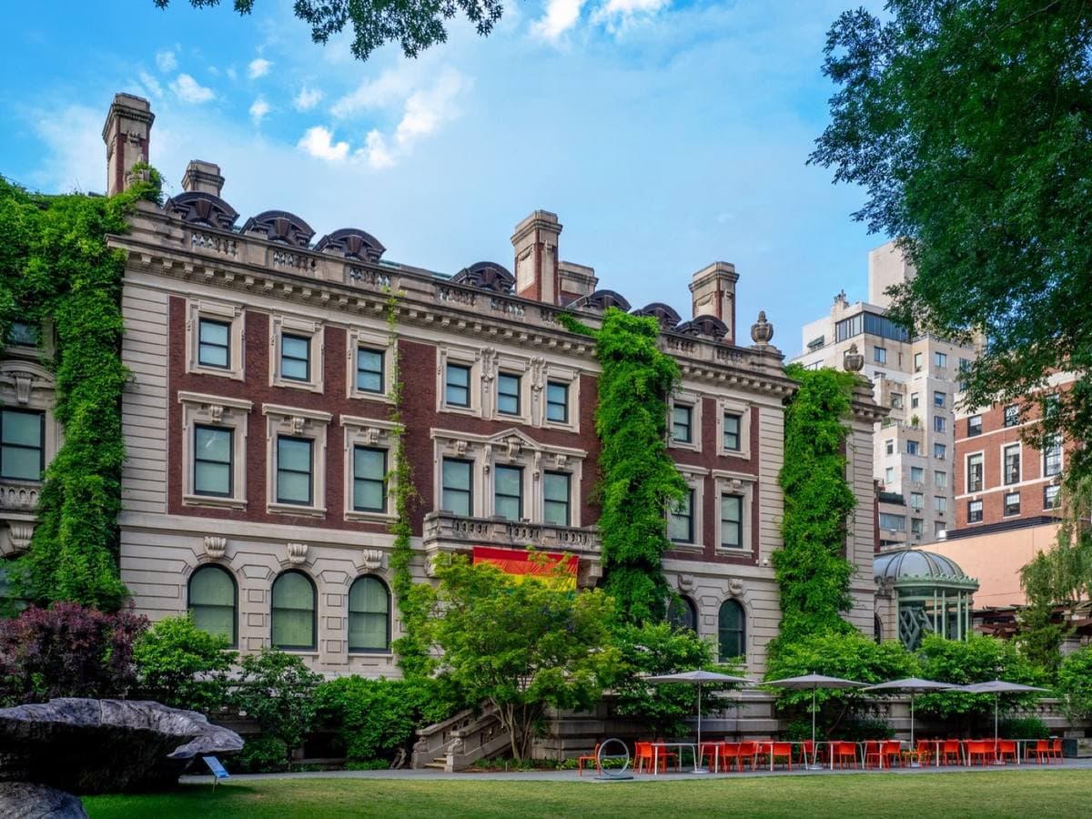 A red-brick mansion with ivy, a garden and red chairs in front