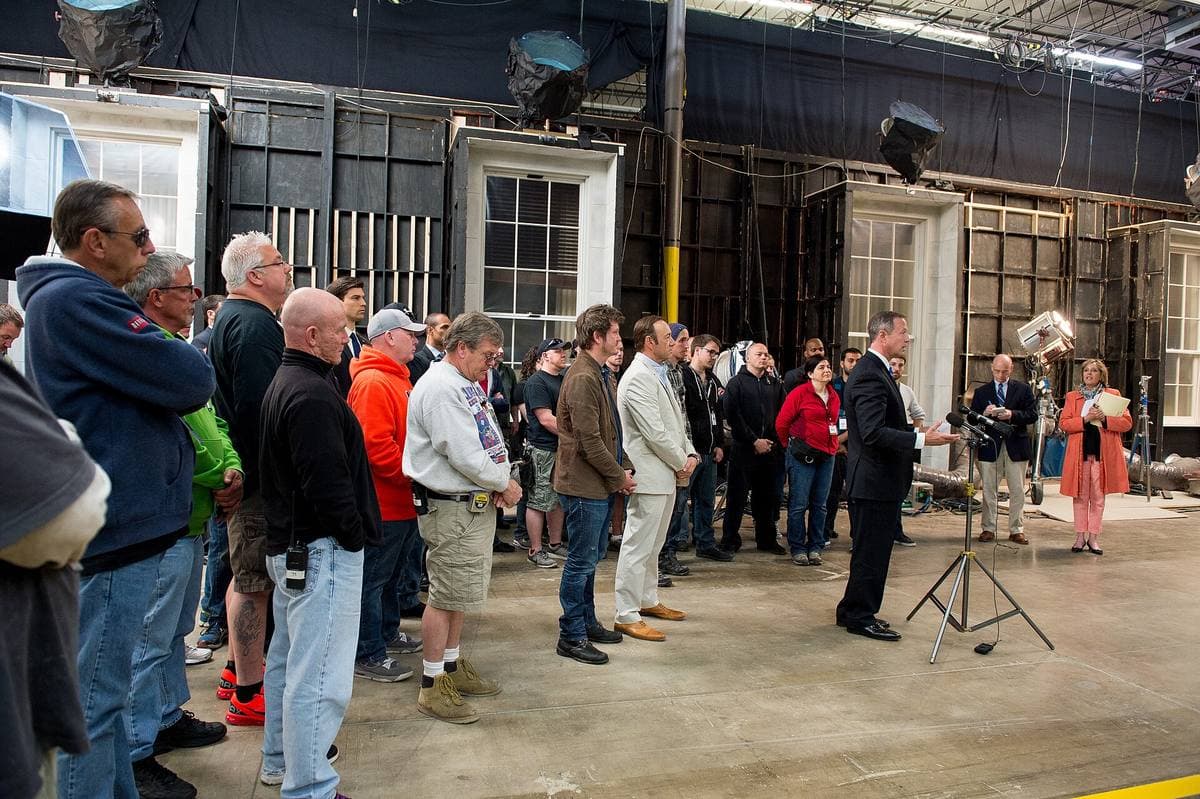 A large crew standing on a soundstage floor in front of built set walls with tall windows