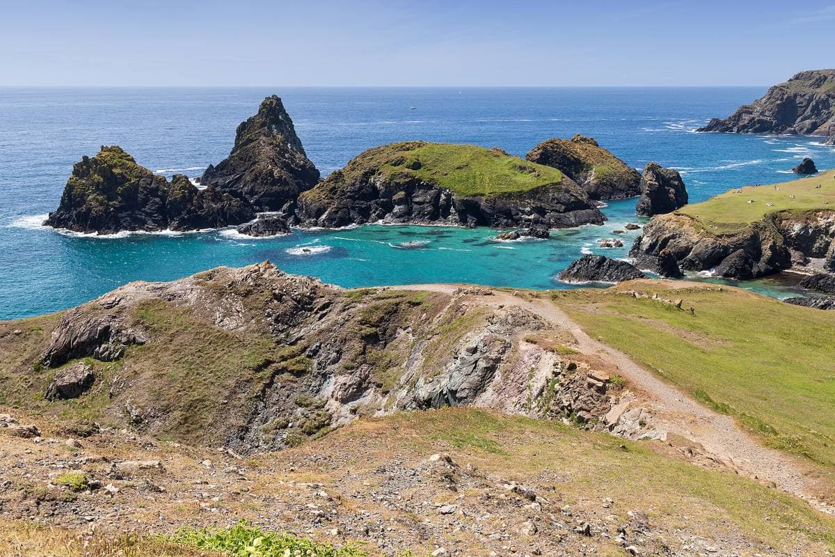Turquoise water around dark rock stacks and a green island, seen from a rocky path