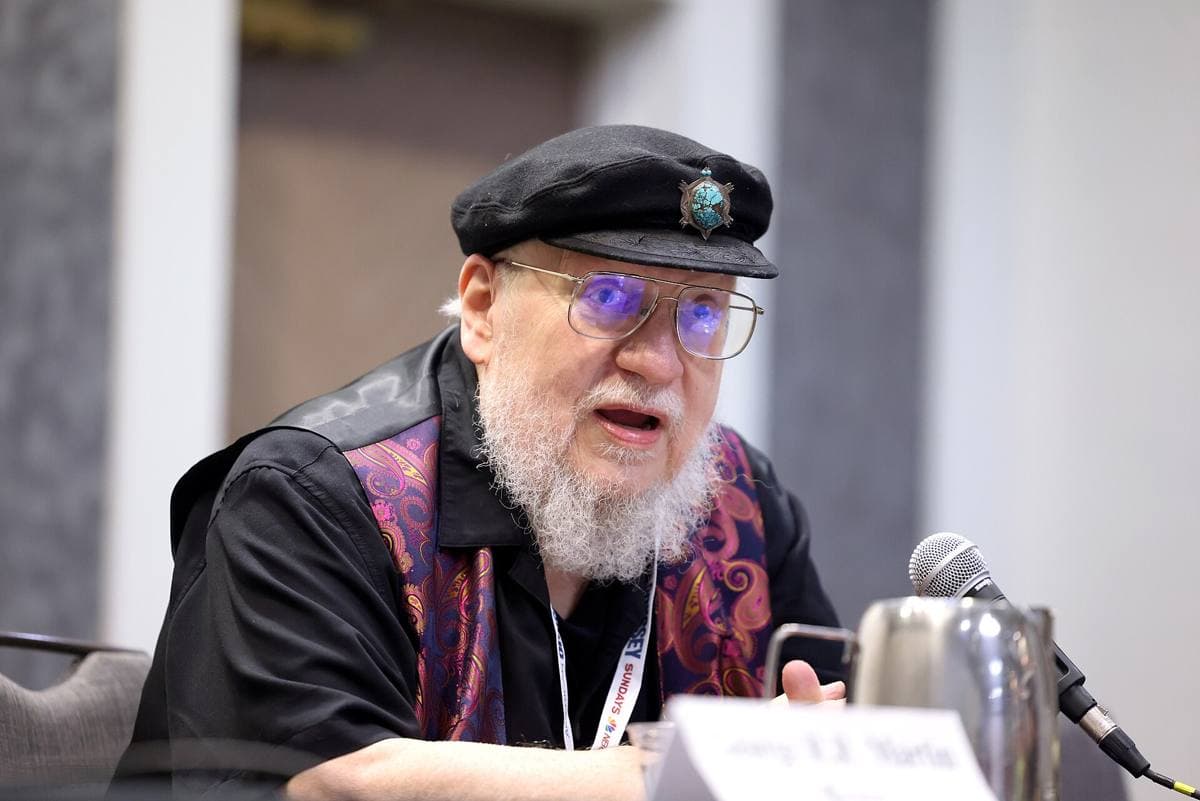 An older man with a white beard and a black cap talking with people at a table