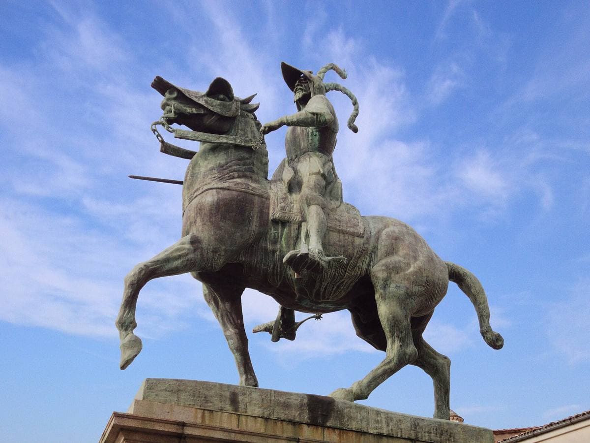 A bronze statue of a helmeted rider on a horse on a stone plinth, a church tower behind