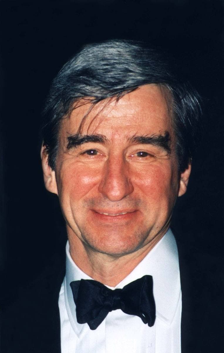 A grey-haired man in a dinner jacket and black bow tie, smiling, against a black background