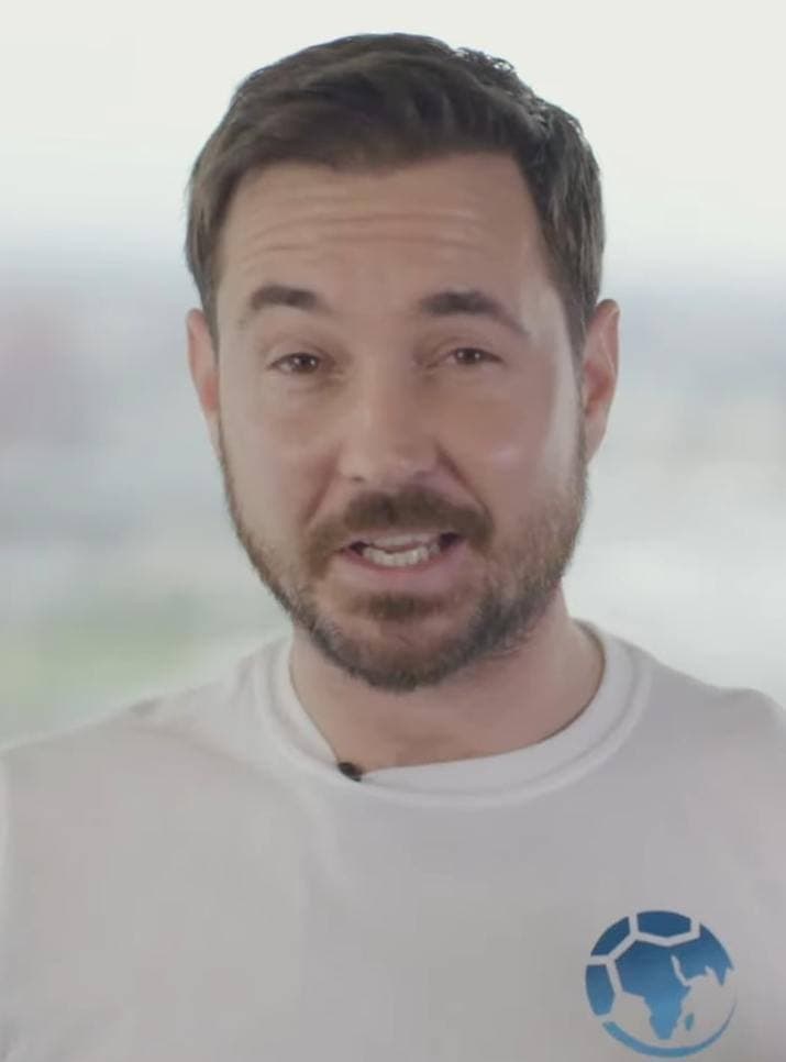 A man with short dark hair and a beard talking to camera in a white T-shirt