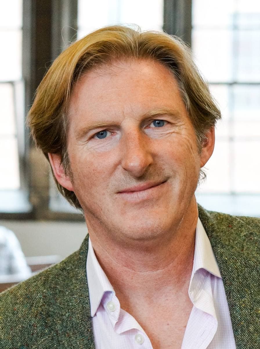 A man with swept-back fair hair smiling in a green jacket and open-necked shirt