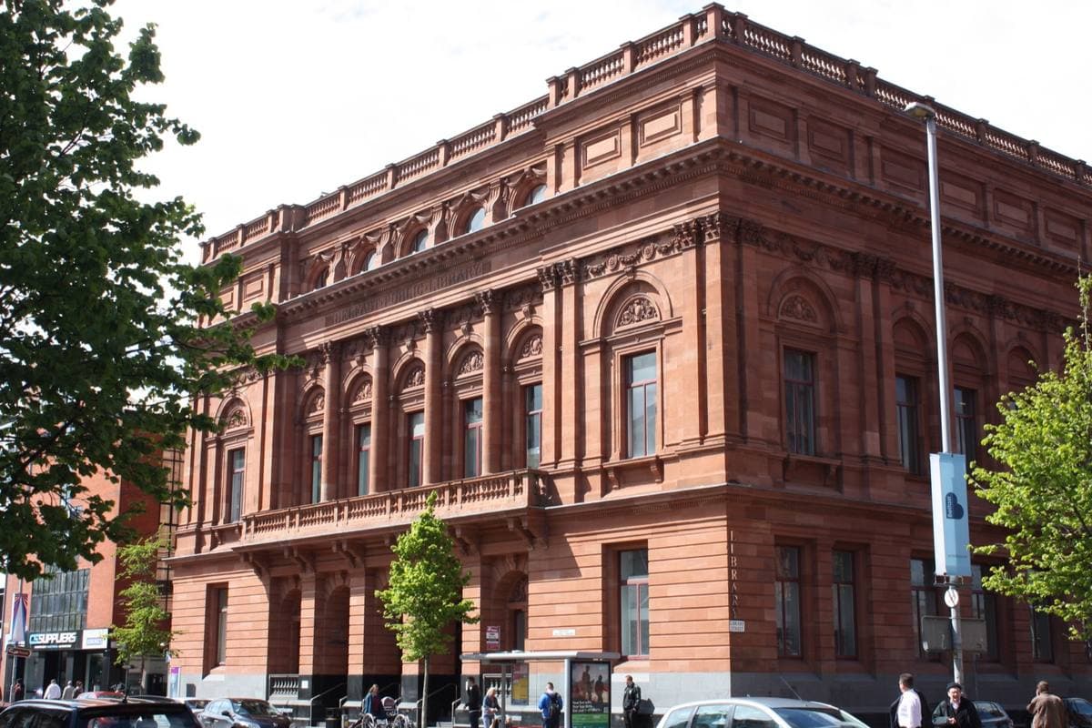 A long red sandstone building with tall arched windows and a heavy cornice on a city street