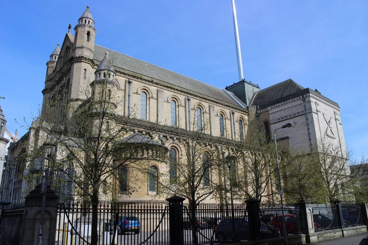 The pale stone west front of a cathedral with three round-arched doorways and a tall steel spire behind