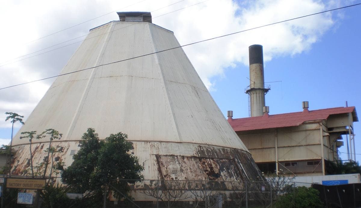 A tall conical grey mill building and a smokestack beside a red-roofed shed