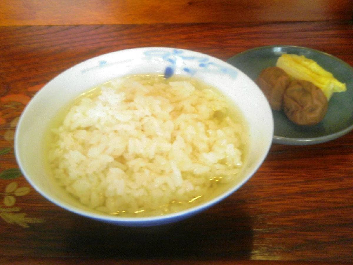 A bowl of rice in pale tea with a small dish of pickled plum beside it