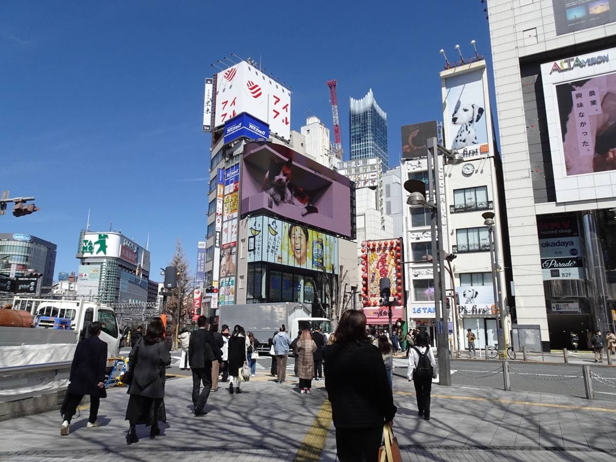 Crowds crossing the plaza on the east side of Shinjuku Station under big screens and shop signs