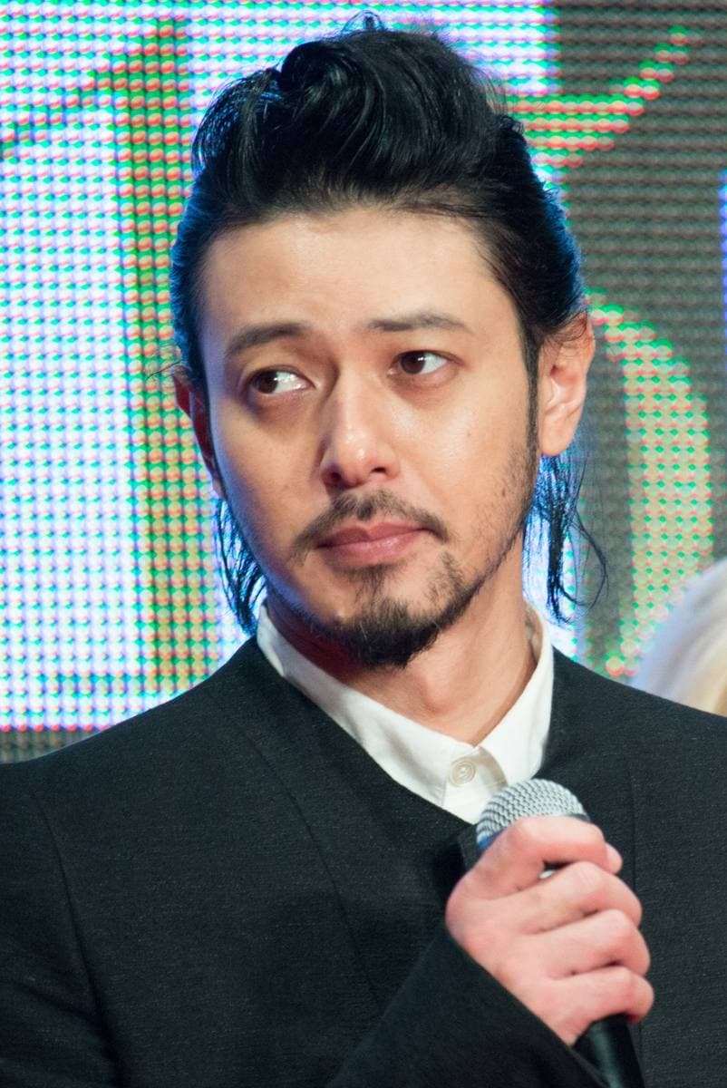 Joe Odagiri in a dark jacket with swept-back hair, holding a microphone