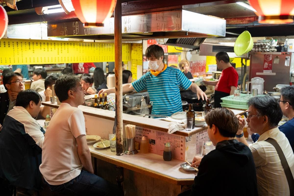 Diners on stools at a small counter in Omoide Yokocho, a cook working behind it under red lanterns