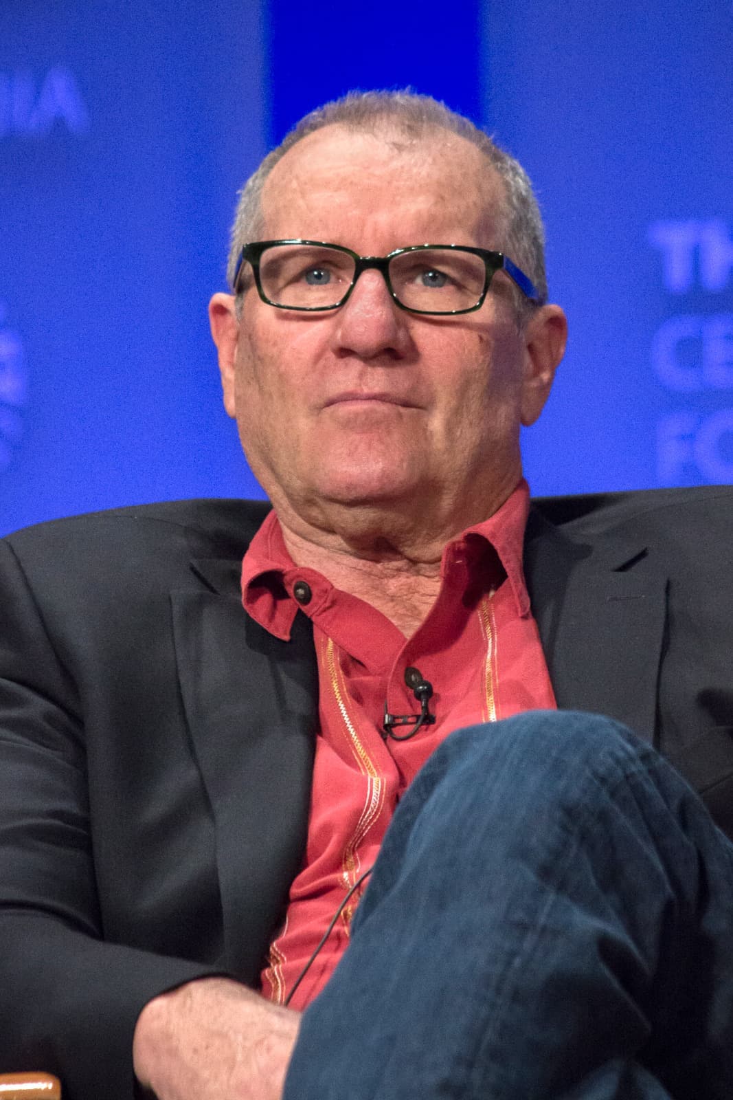 Ed O’Neill in glasses and a red shirt, seated on a panel stage