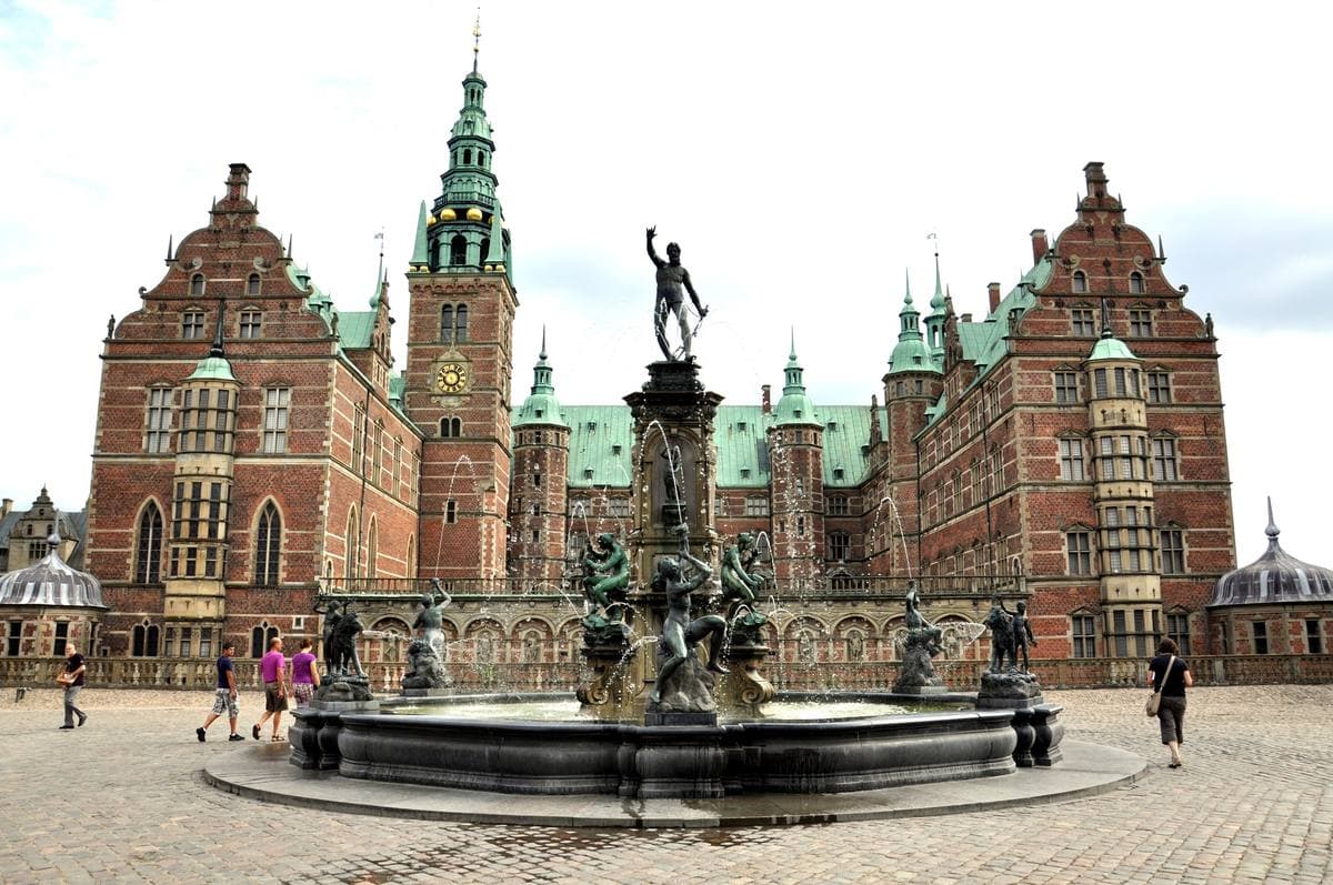 A red brick Renaissance castle with green spires reflected in a lake