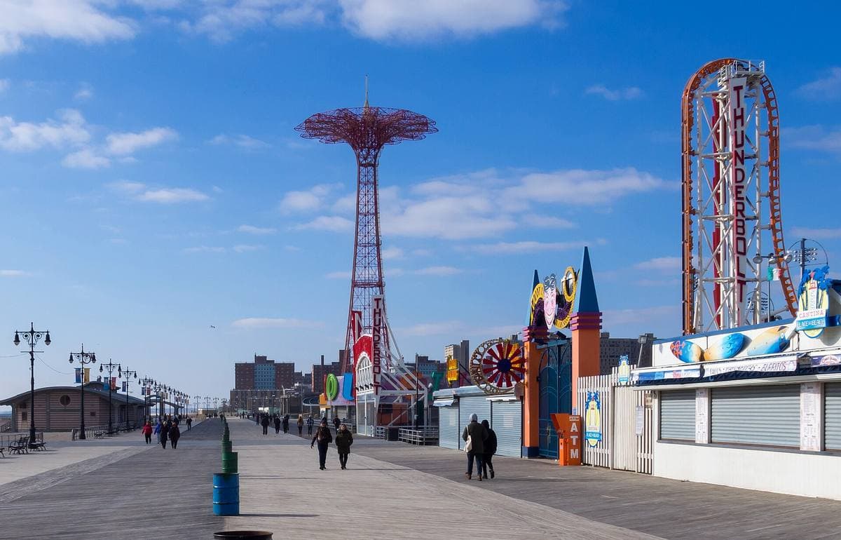 A wide wooden boardwalk with the Parachute Jump and a roller coaster in the distance