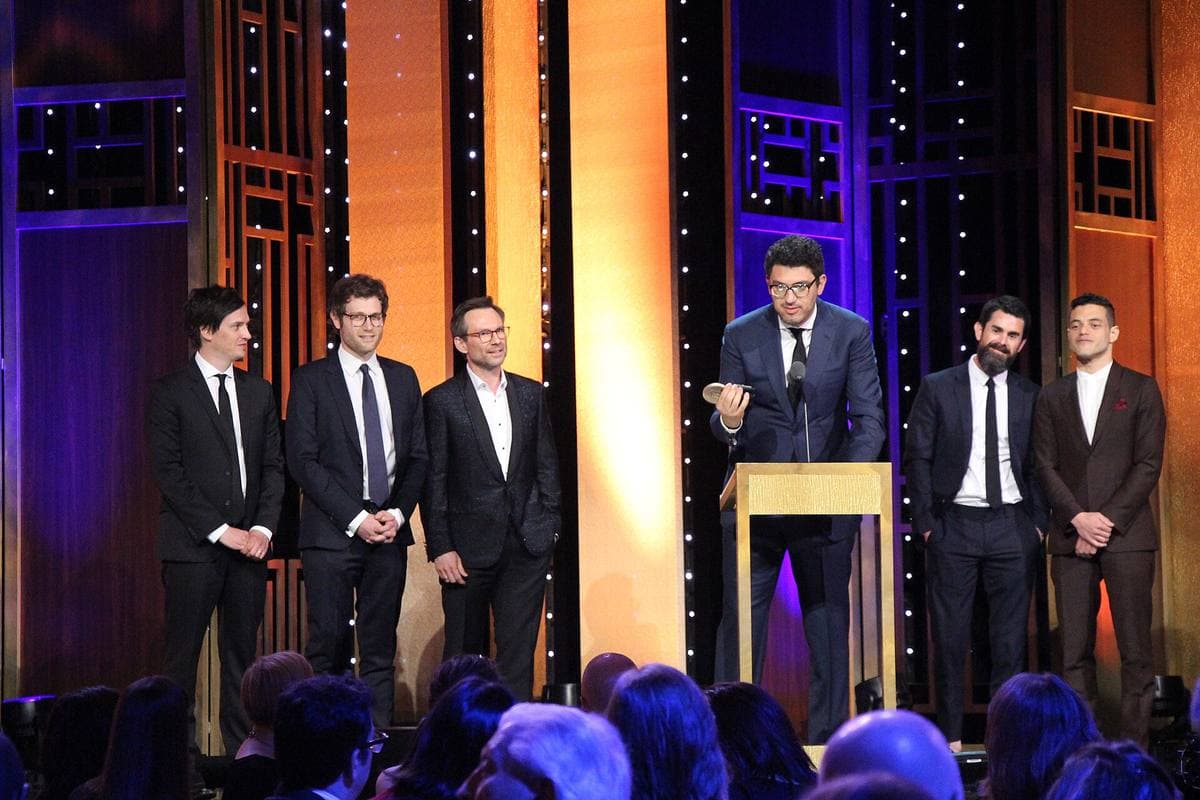 Six men in dark suits on a stage at a lectern