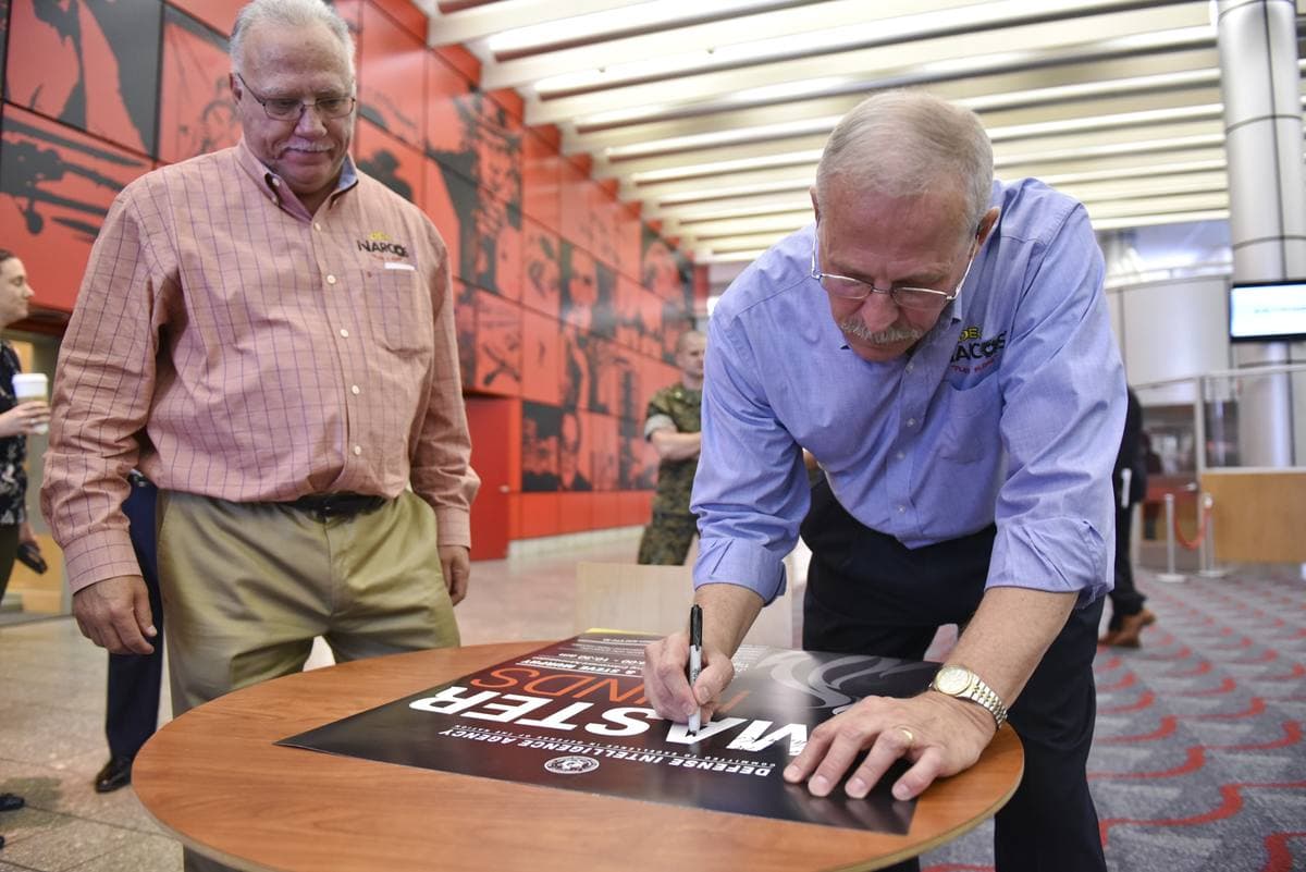 Two older men at a table, one signing a poster while the other looks on