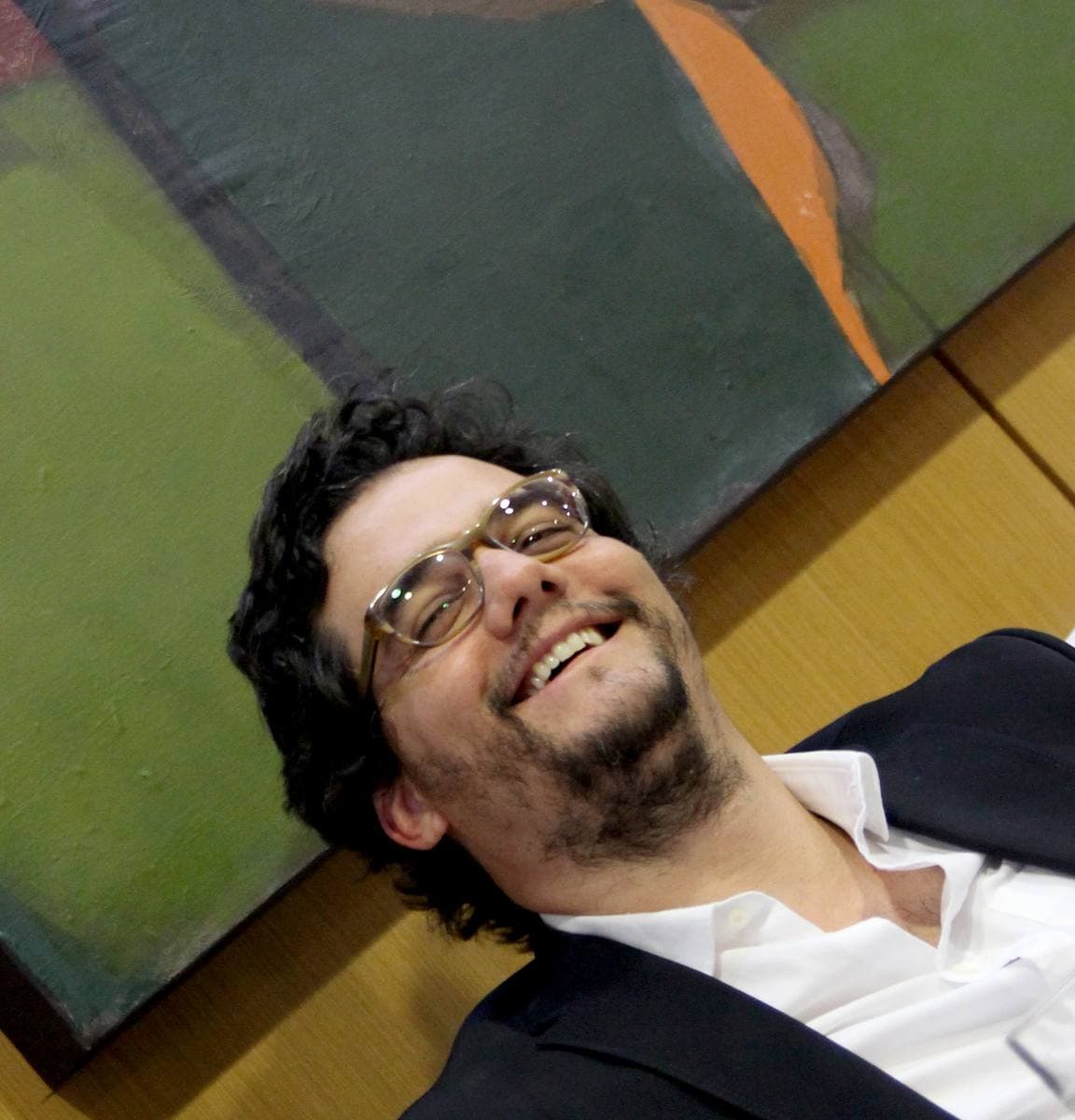 A smiling man with dark curly hair, glasses and a beard, leaning back