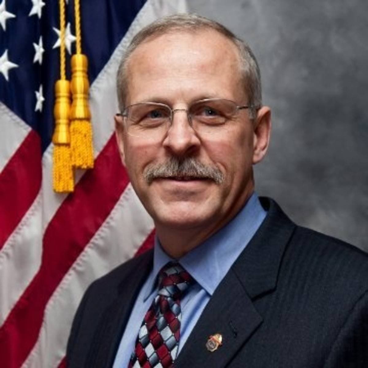 A grey-haired man with glasses and a moustache in a suit in front of a flag