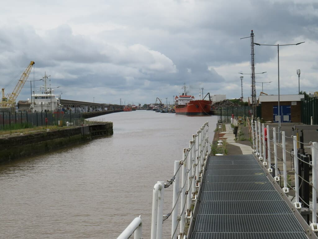 A walkway beside a dock with a red ship and cranes beyond