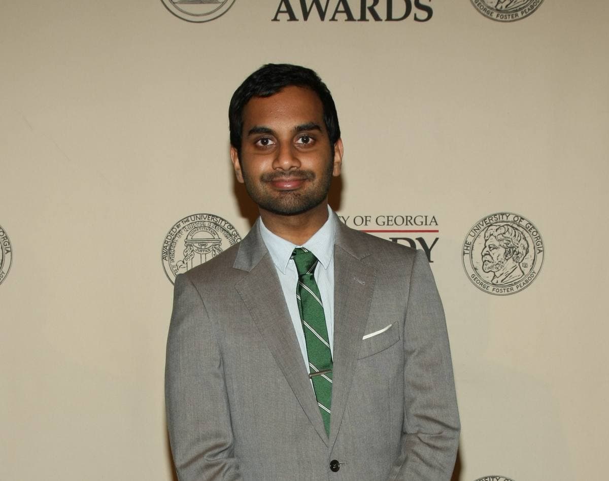 Aziz Ansari in a grey suit and green striped tie against a Peabody Awards backdrop
