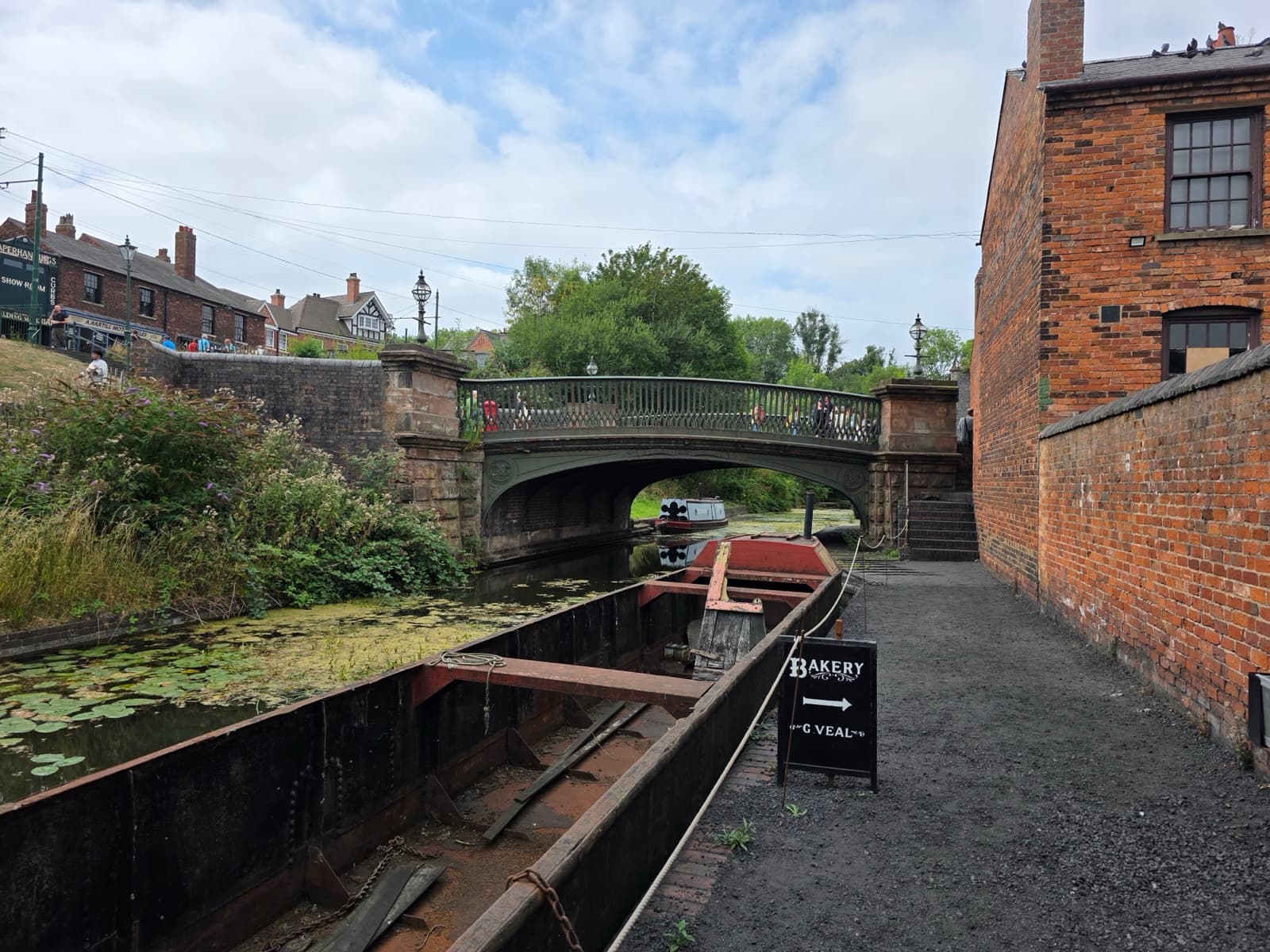 A narrow canal with a moored boat, a brick bridge and a towpath beside brick workshops